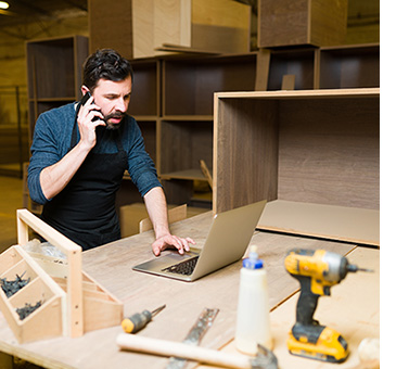 Cabinet maker in his shop on the phone and in front of a laptop